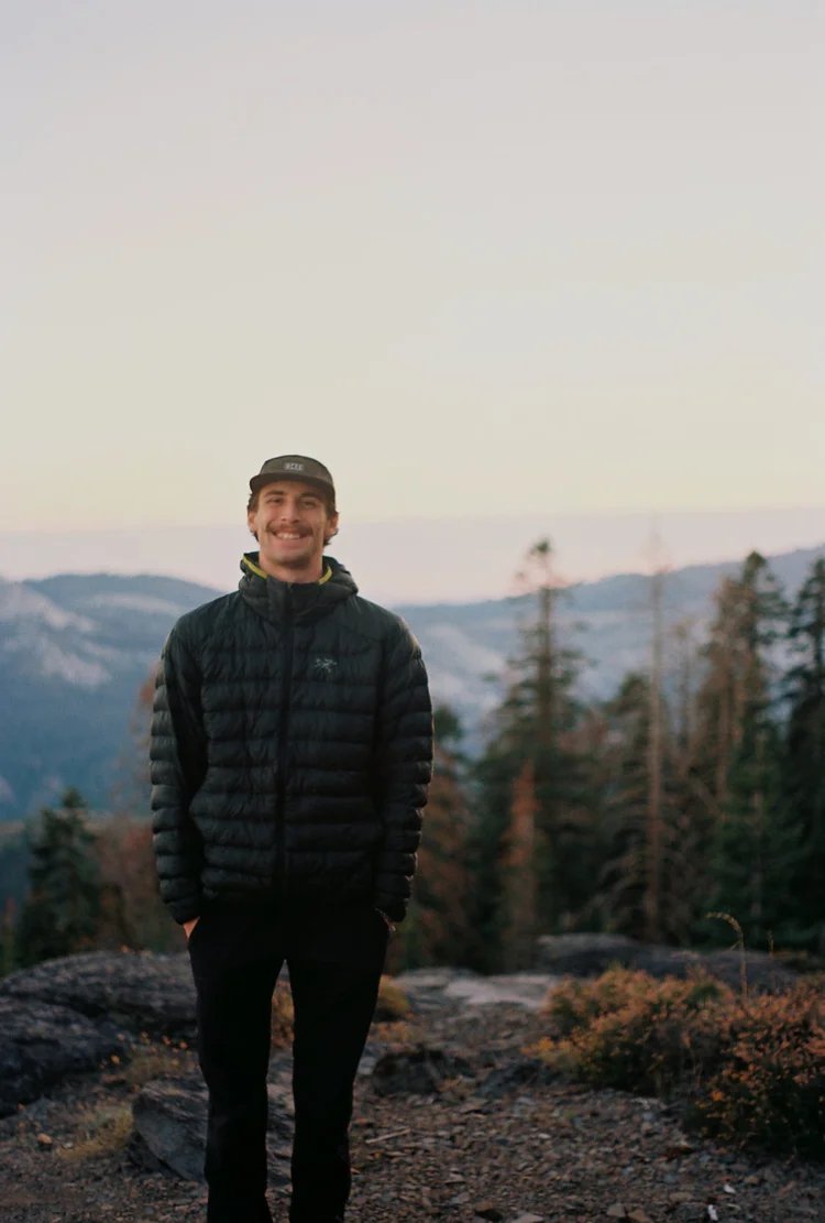 Moses Wagner smiling outdoors in the mountains, wearing a cap and puffer jacket